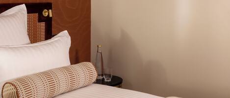 A neatly made bed with a rolled-up blanket, a bedside table with a bottle and glasses, and framed artwork on the wall.