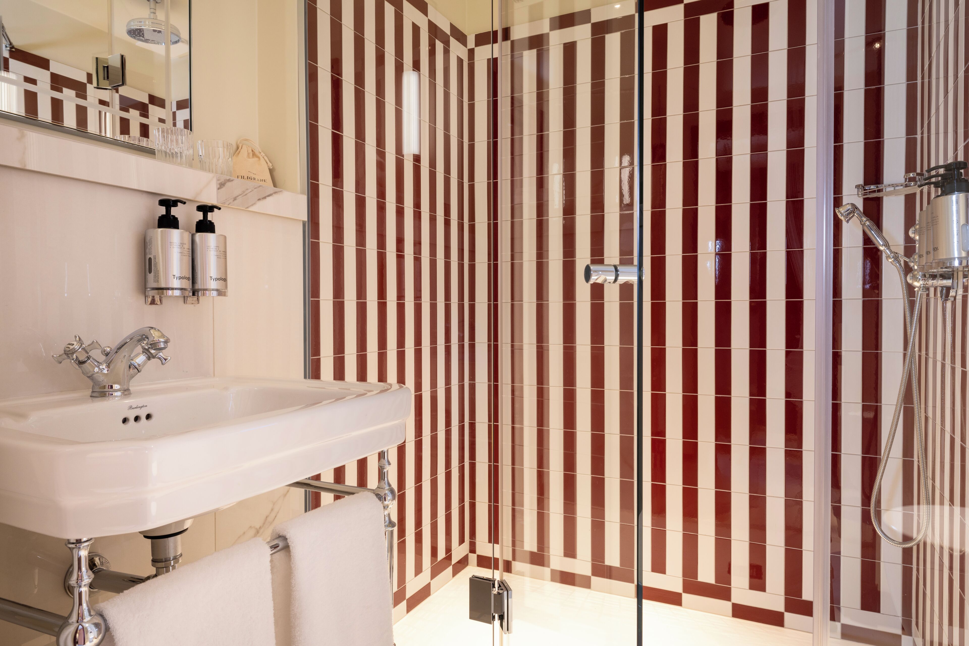 A bathroom with a white sink, a mirror, two bottles of Typology, a shower with a handheld showerhead, and a striped wall.
