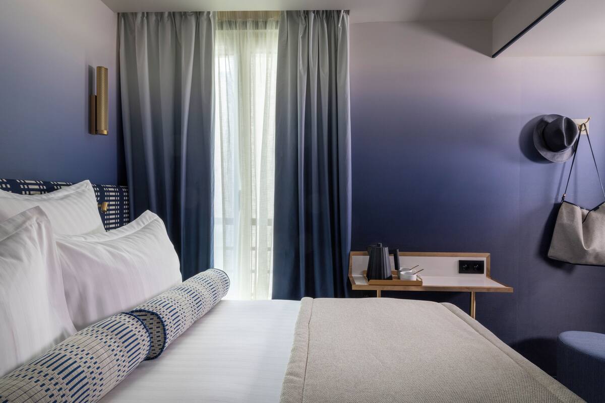 Una habitación de hotel con una cama, una mesita de noche con lámpara, una silla y una ventana con cortinas.