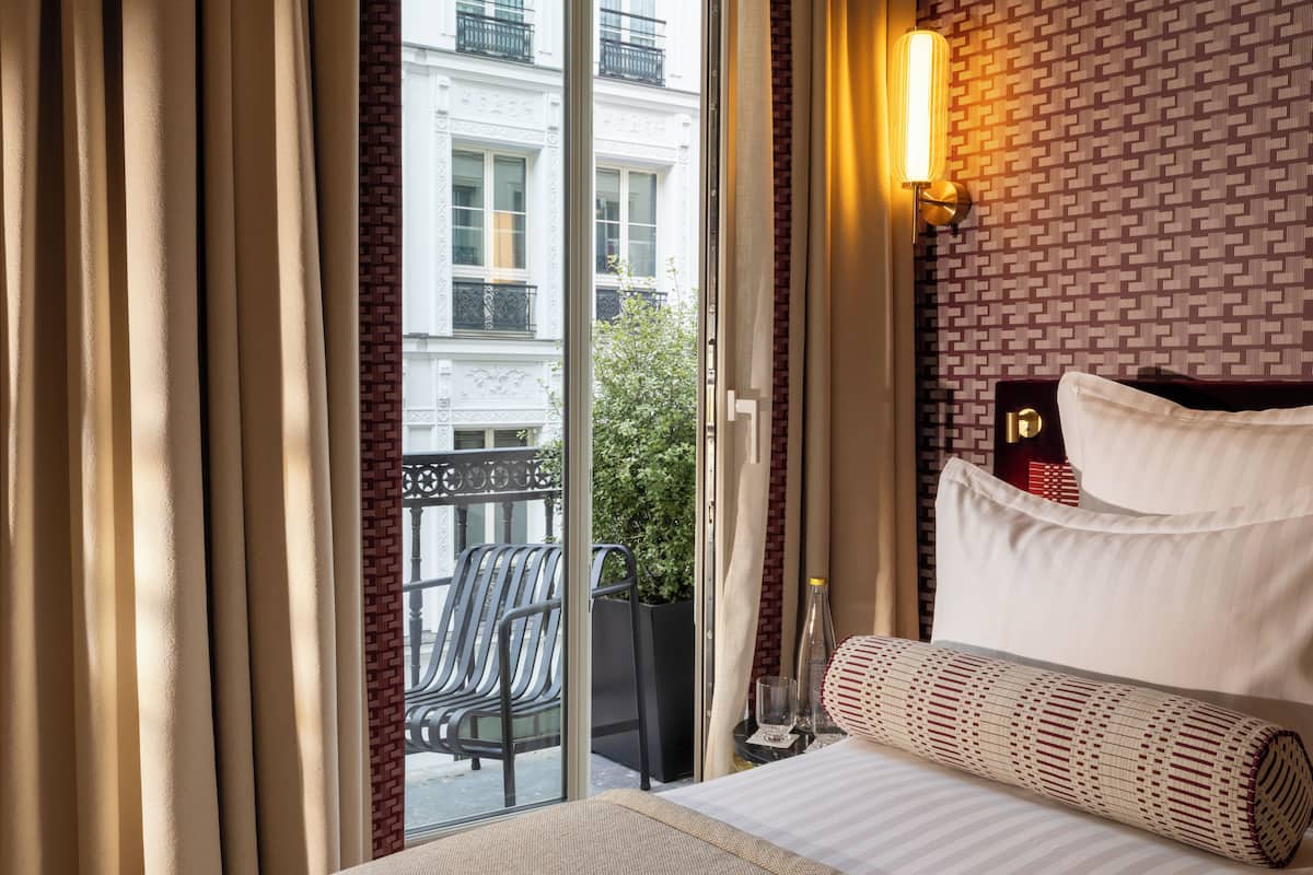 Habitación de hotel con cama, cortinas, lámpara de pared y vistas al balcón exterior.