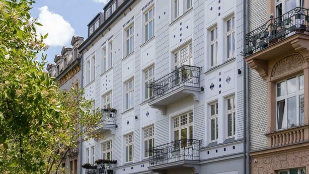 Une rangée d’immeubles à plusieurs étages dotés de balcons, des voitures garées et un ciel dégagé.