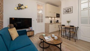 Un salon moderne avec un canapé bleu, un téléviseur à écran plat, une table basse avec des magazines, un coin repas avec une table et des chaises en bois, et une cuisine avec des meubles blancs et un four à micro-ondes.