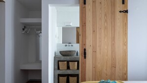 A bedroom with a wooden door, a bed with a blue blanket, a bathroom with a white sink and a mirror, and a shelf with baskets.
