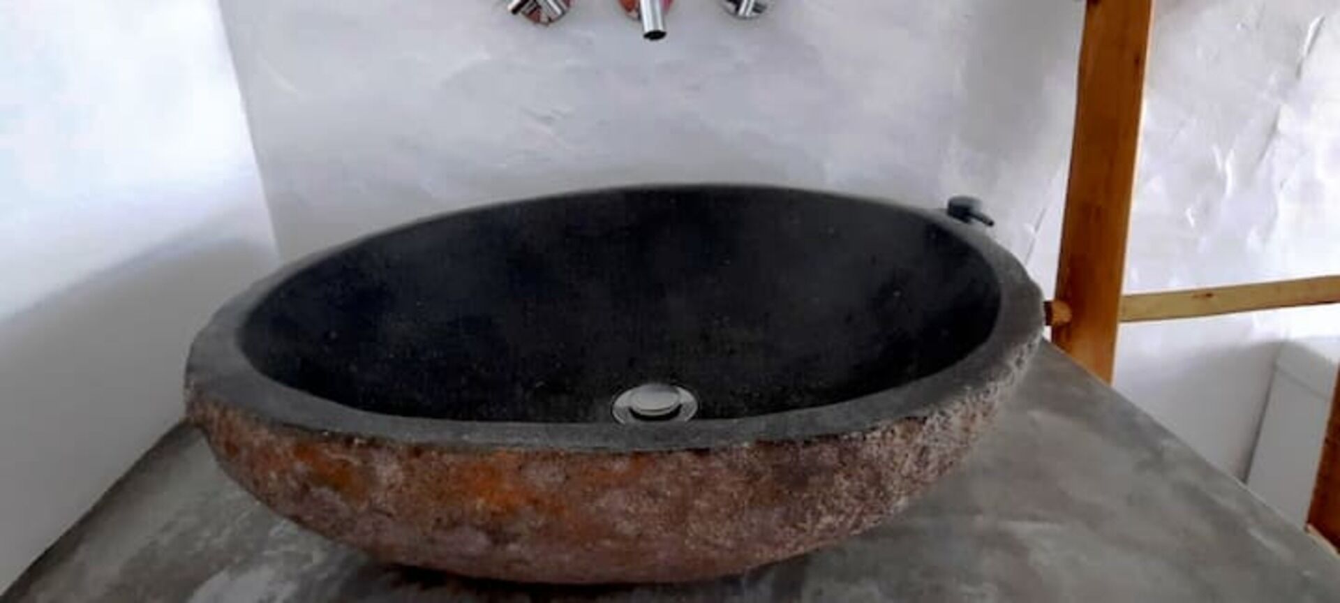 Un lavabo de piedra rústico y redondo, con desagüe metálico, frente a una pared lisa.