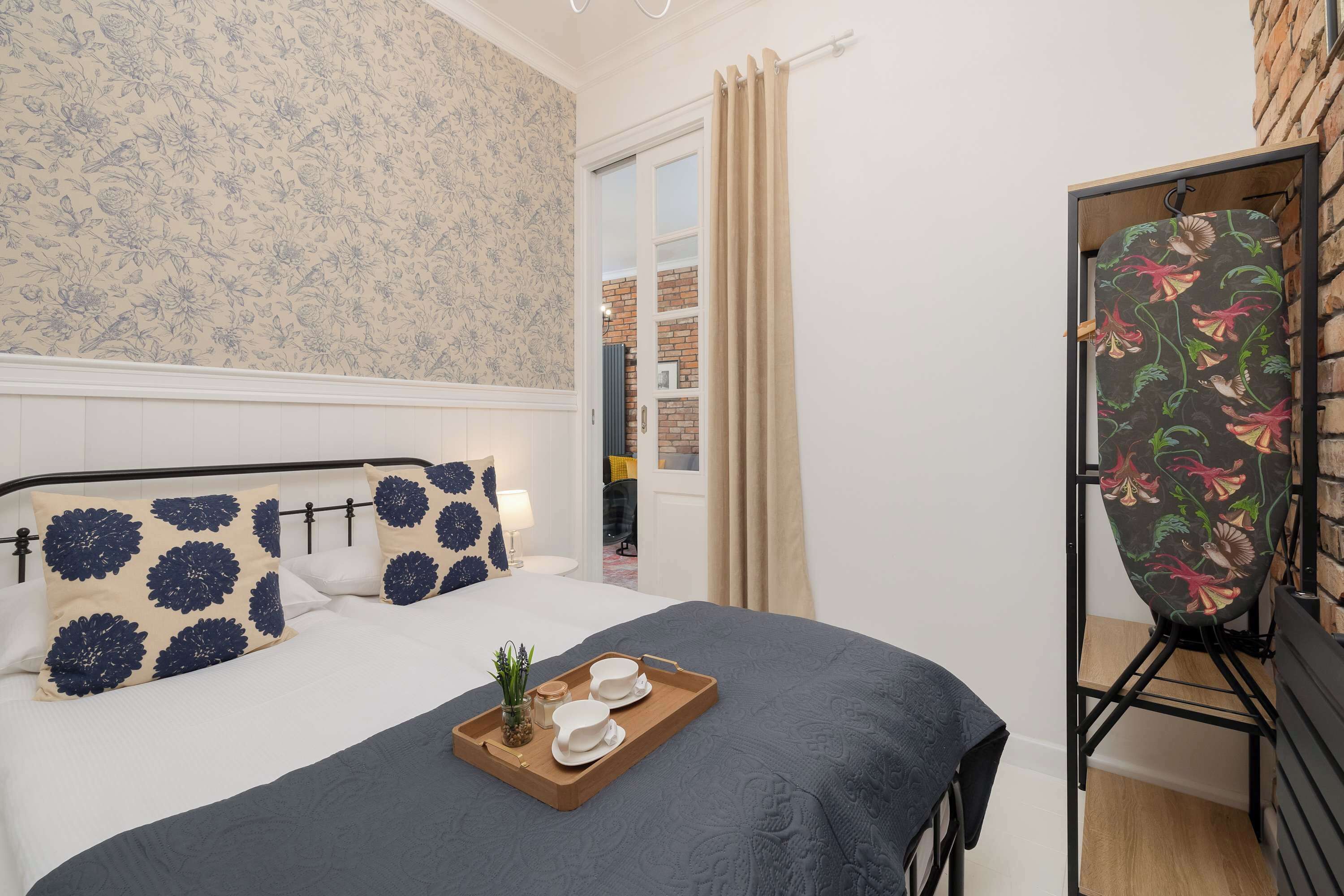 Ein ordentlich bezogenes Bett mit einem Tablett mit Frühstücksgebäck, eine Wand mit Blumenmuster und Blick auf ein Backsteingebäude durch eine offene Tür.
