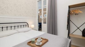 A neatly made bed with a tray of tea and a candle, a wooden shelf with clothes, and a view of a brick wall through an open door. - Stylish Apartment | Balcony and Air Conditioning (Kraków)
