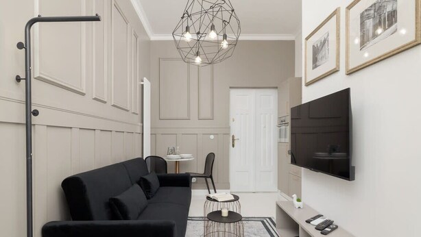 A modern living room with a black sofa, a TV mounted on the wall, and a geometric light fixture. - Tasteful Apartment with Balcony | Air Conditioning | Close to the Center (Kraków)