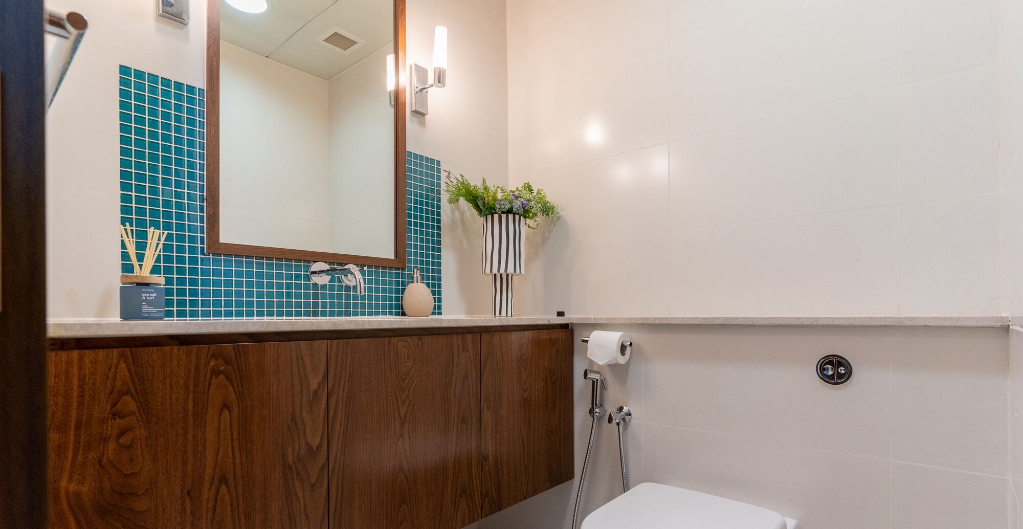 Ein Badezimmer mit einem Holzkosmetikschrank, einem Spiegel, einer blau gefliesten Wand, einer Toilette und einer Pflanze auf der Ablagefläche.
