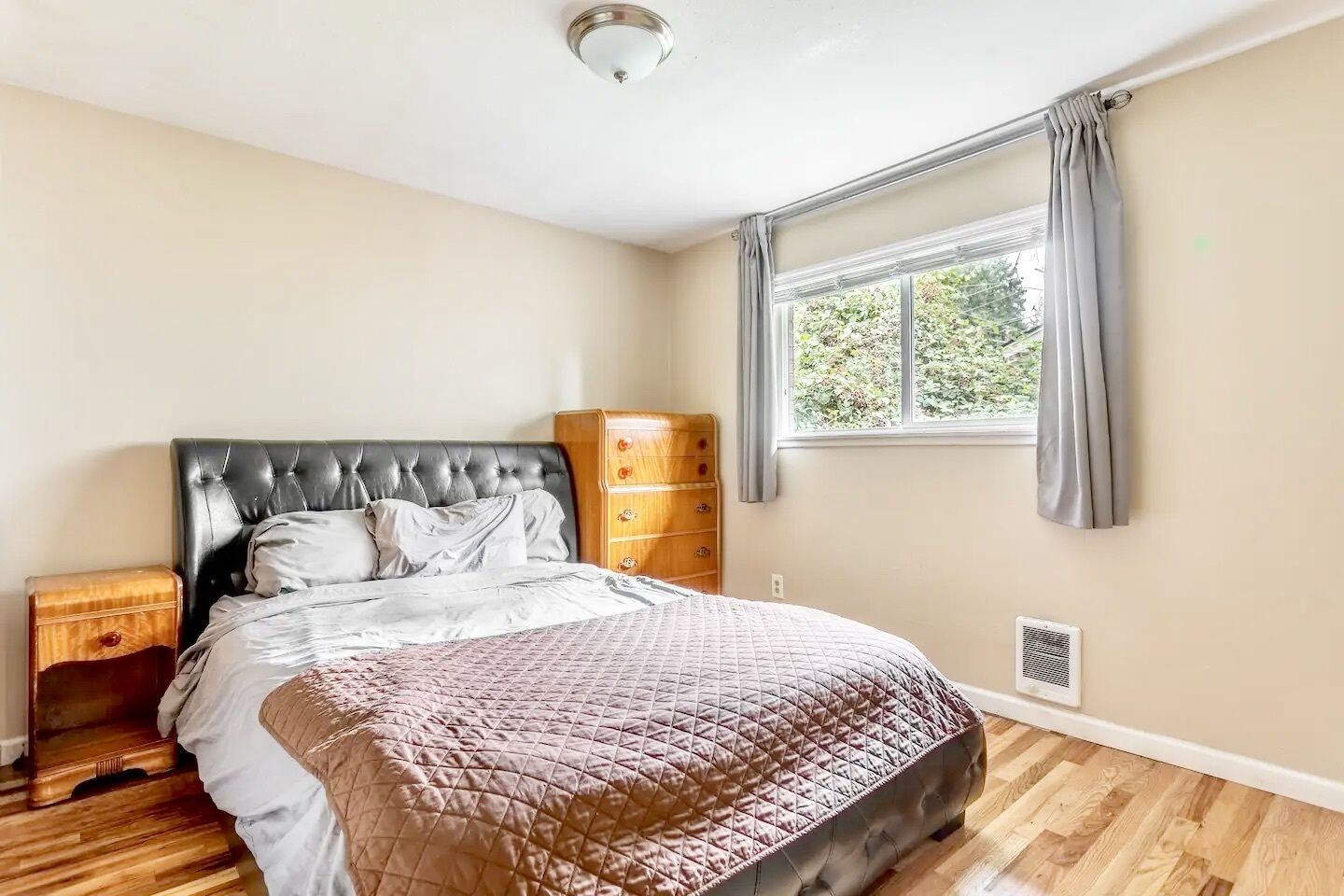 A bedroom with a bed, wooden nightstand, dresser, window with curtains, and a ceiling light.