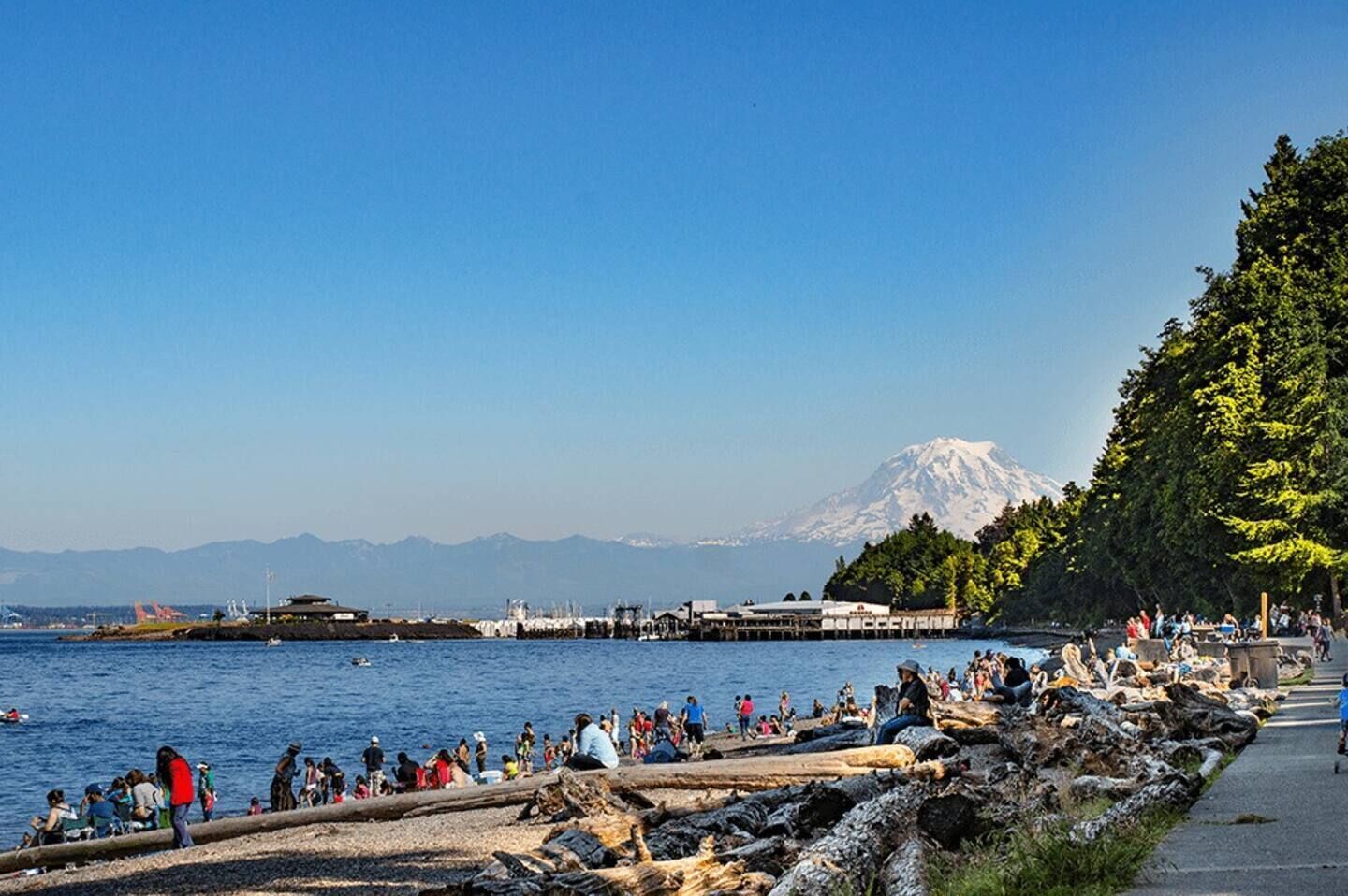 A coastal area with a rocky shoreline, a clear view of a mountain in the background, and a paved walkway along the water's edge.