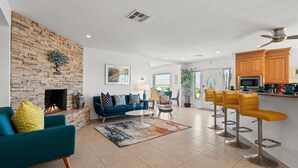 A spacious living room with a fireplace, a blue sofa, a coffee table, and a kitchen area with wooden cabinets and bar stools.