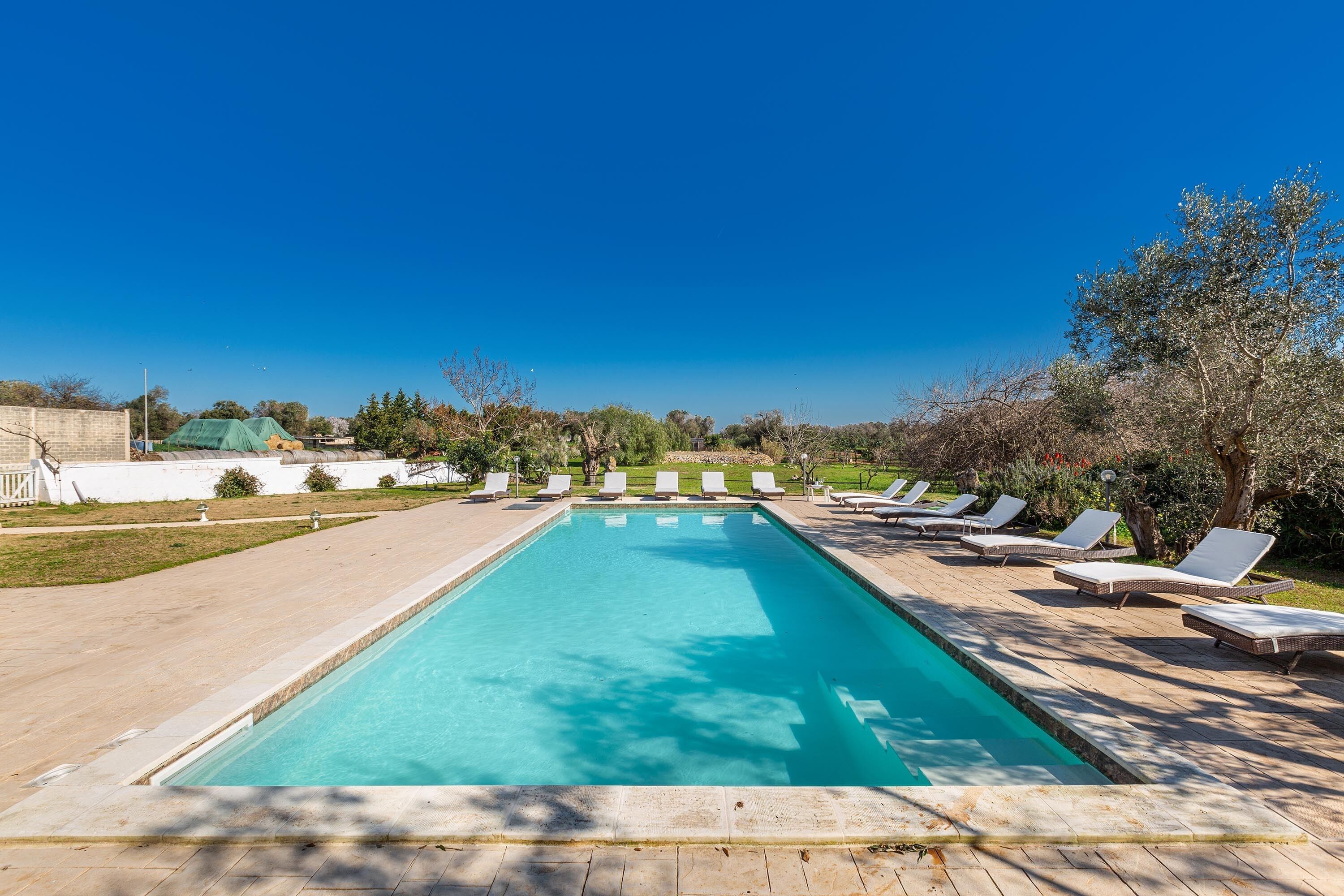 A rectangular swimming pool with a tiled deck, surrounded by white lounge chairs and a grassy area with trees and shrubs.
