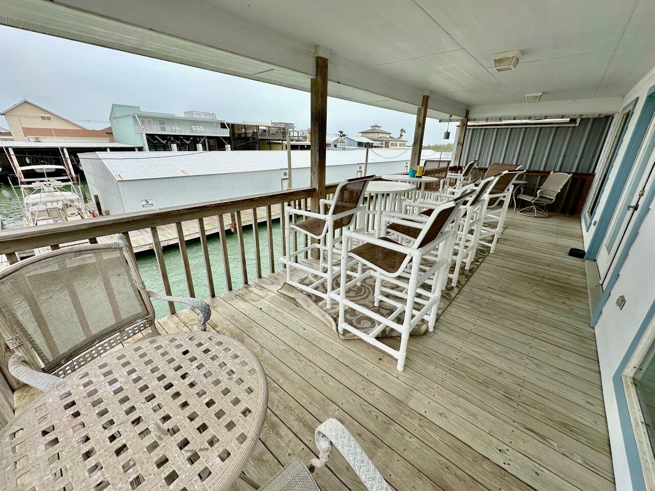 Une terrasse extérieure couverte, dotée de chaises et d’une table en osier blanc, qui offre une vue sur une marina avec des bateaux et des bâtiments.