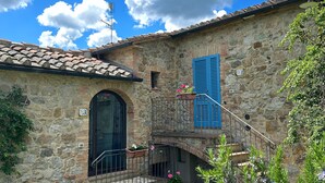 A stone building with a tiled roof, a staircase leading to a balcony, and a small garden area with flowers. - Borgo Tignano (Volterra)