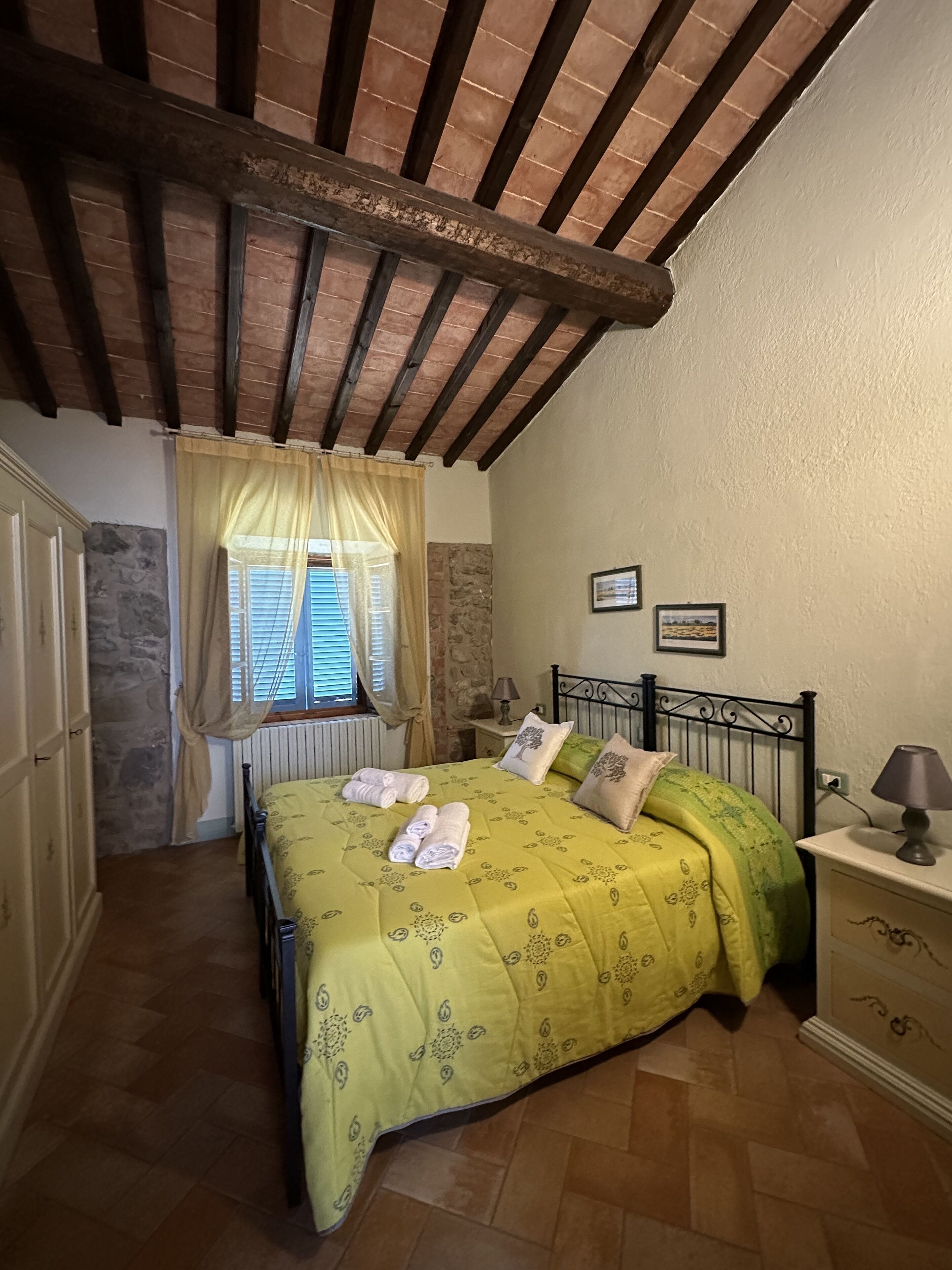 Una camera da letto con un letto, un comodino, una finestra con tende, un muro in pietra e un soffitto in legno.