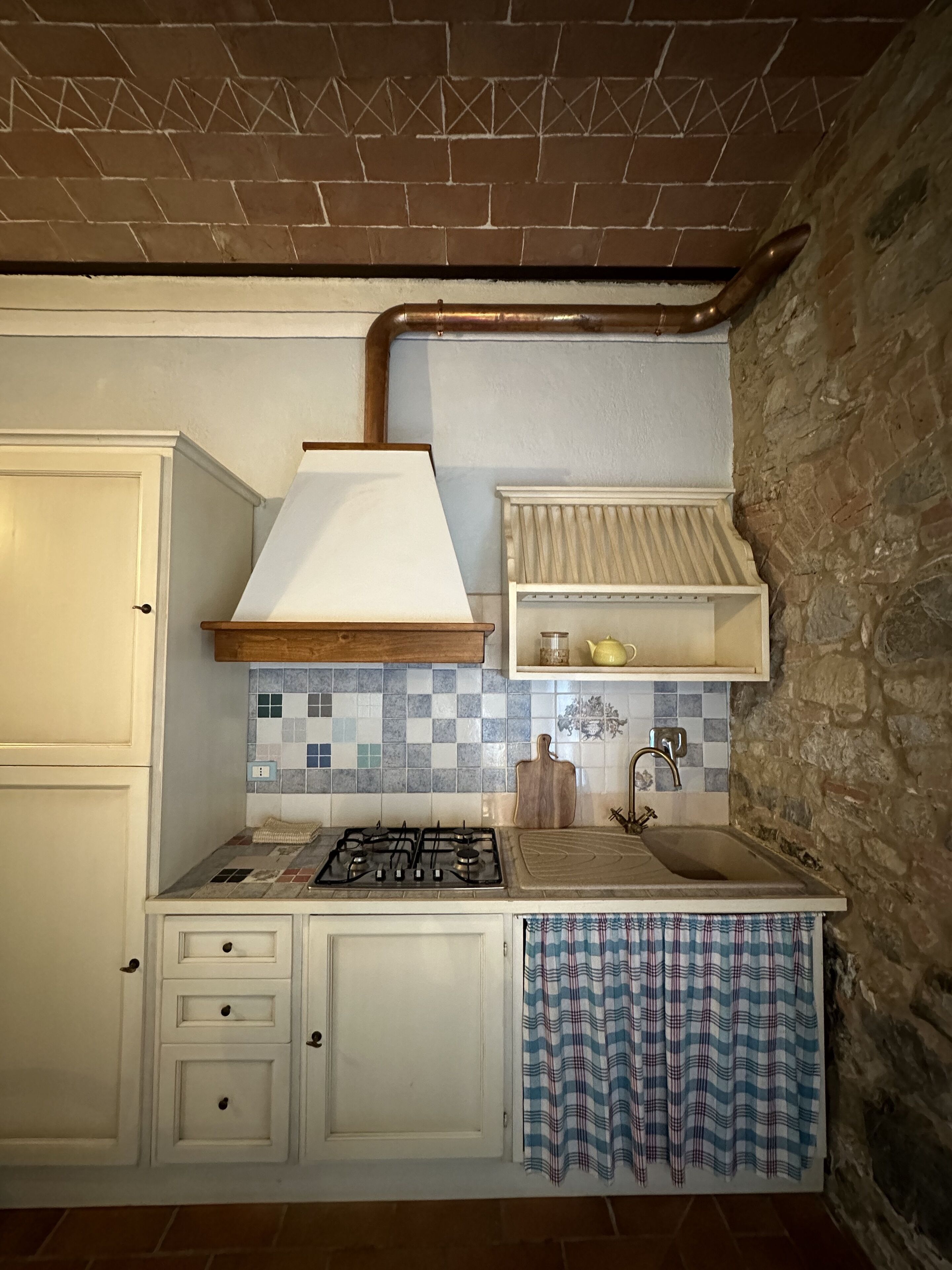 Una cocina compacta con estufa a gas, armarios blancos, salpicadero de azulejos y pared de piedra.