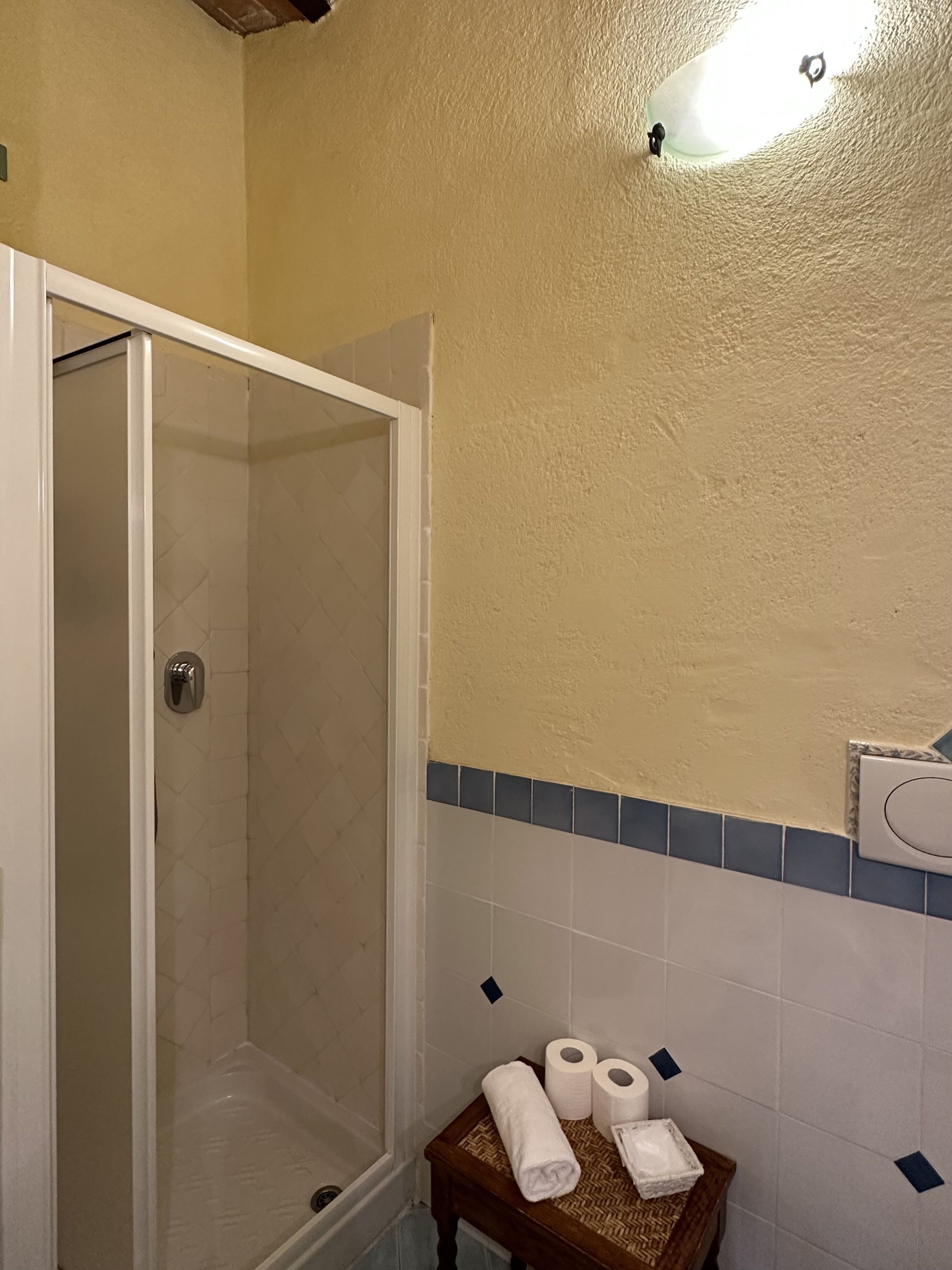 A bathroom with a glass shower enclosure, a wooden bench with towels, and a tiled wall with a blue accent strip.