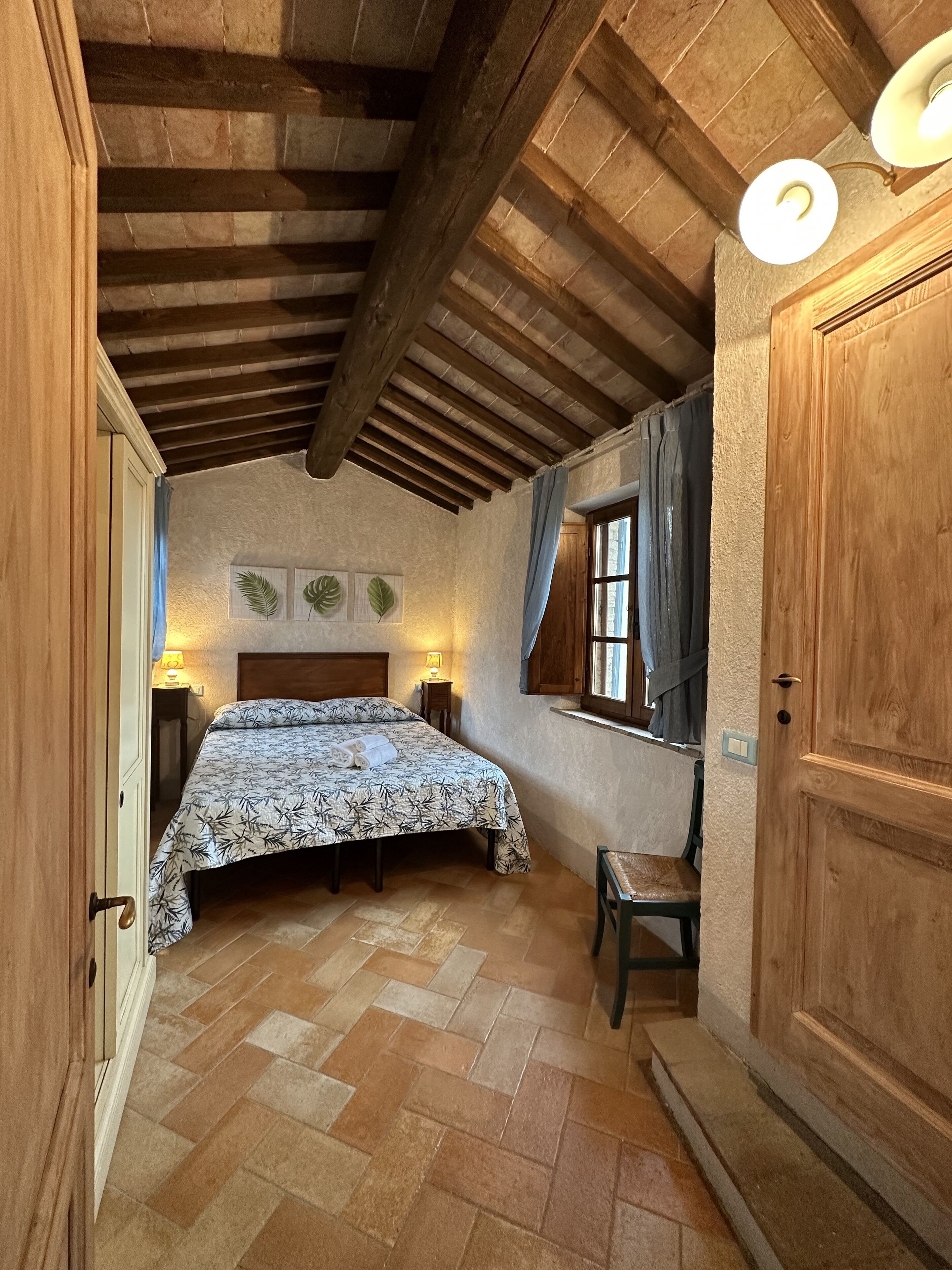 Una camera da letto con soffitto in legno, un letto con copriletto decorato, un comodino con lampada e una finestra con tende.