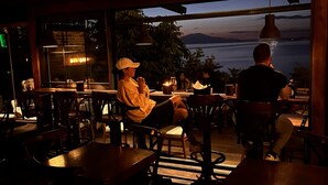 Un comedor interior con mesas y sillas, ventanas amplias con vistas a un cuerpo de agua y lámparas colgantes.