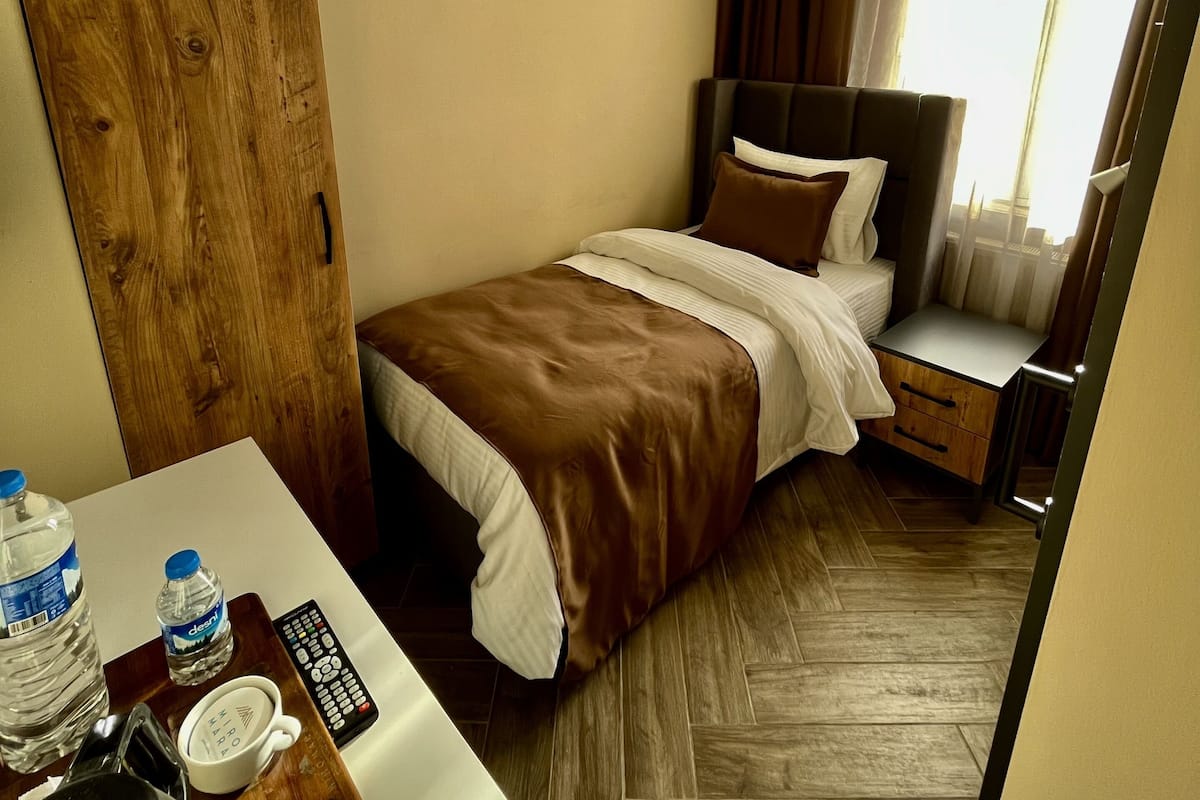 A hotel room with a bed, bedside table, and a desk with a kettle, water bottles, and a cup.