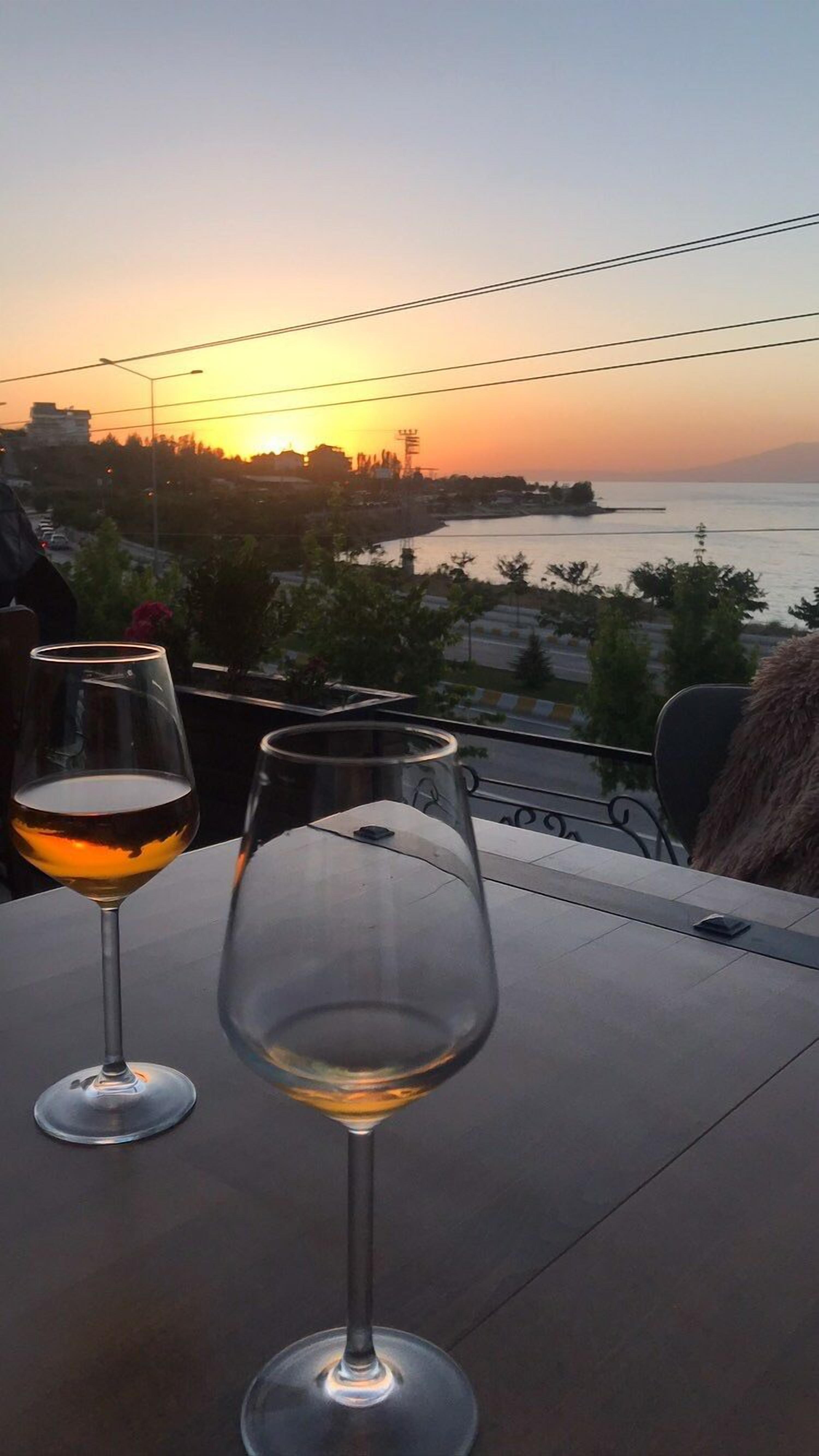 Zwei Weingläser auf einem Tisch mit Blick auf einen Sonnenuntergang über dem Wasser und einer Stadtlandschaft im Hintergrund.