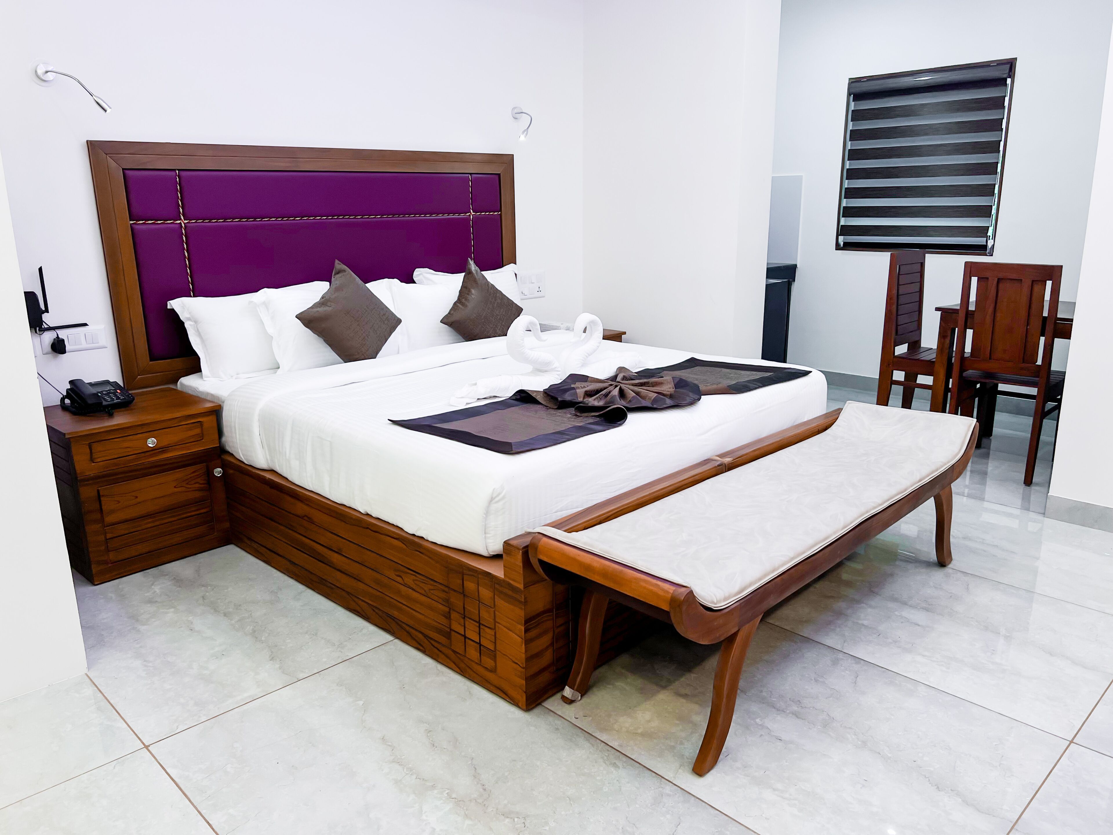 Una cama bien hecha con estructura de madera y cabecera morada, una mesita de noche con un teléfono y un banco al pie de la cama.