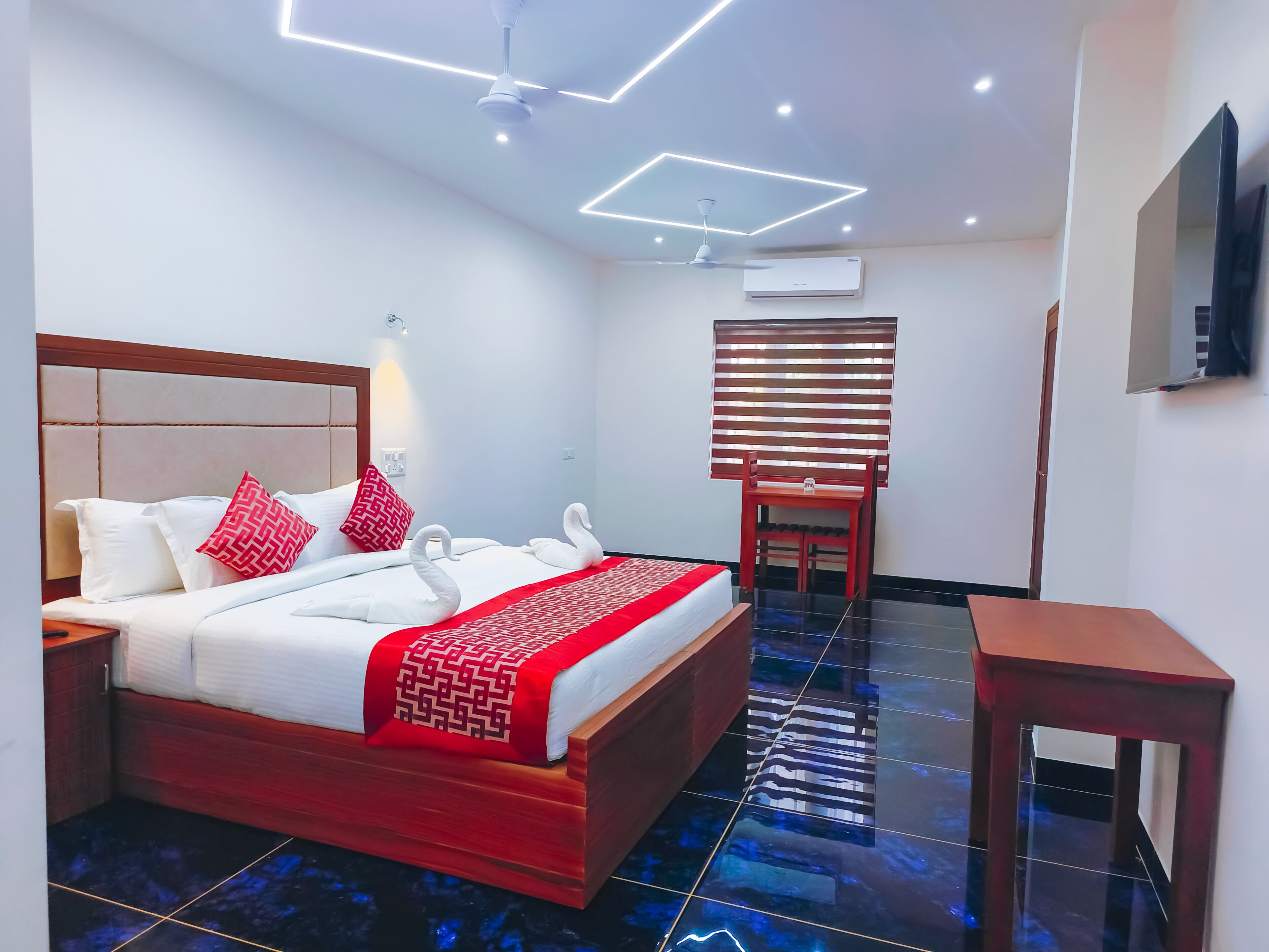 A hotel room with a bed, a wooden nightstand, a red and white patterned bedspread, a red and white pillow, a wooden chair, a small wooden table, a ceiling fan, recessed lighting, and an air conditioning unit.