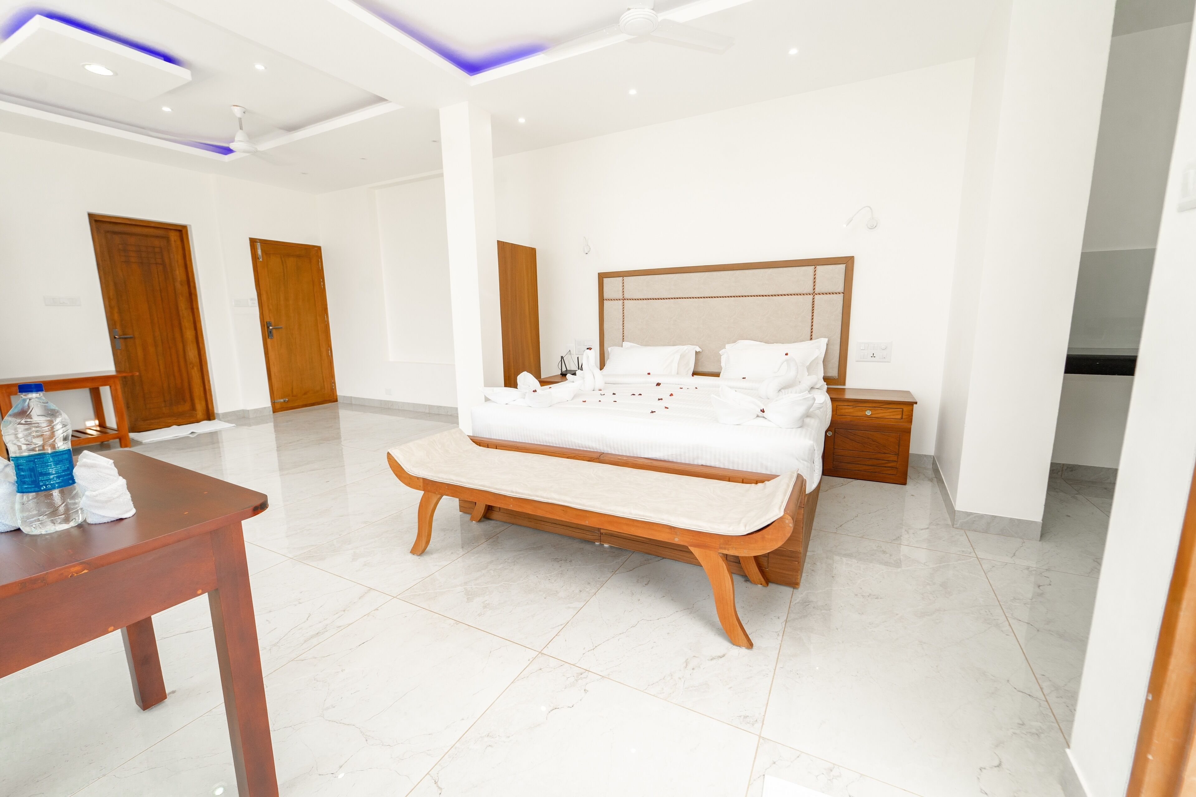 Habitación de hotel moderna con una cama grande, mesitas de noche, un banco de madera y una pequeña mesa con una botella y servilletas.