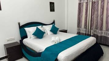A neatly made bed with a teal and white bedspread, two pillows, and a teal blanket. There is a nightstand with a drawer, a wall-mounted light, and a framed picture above the bed.