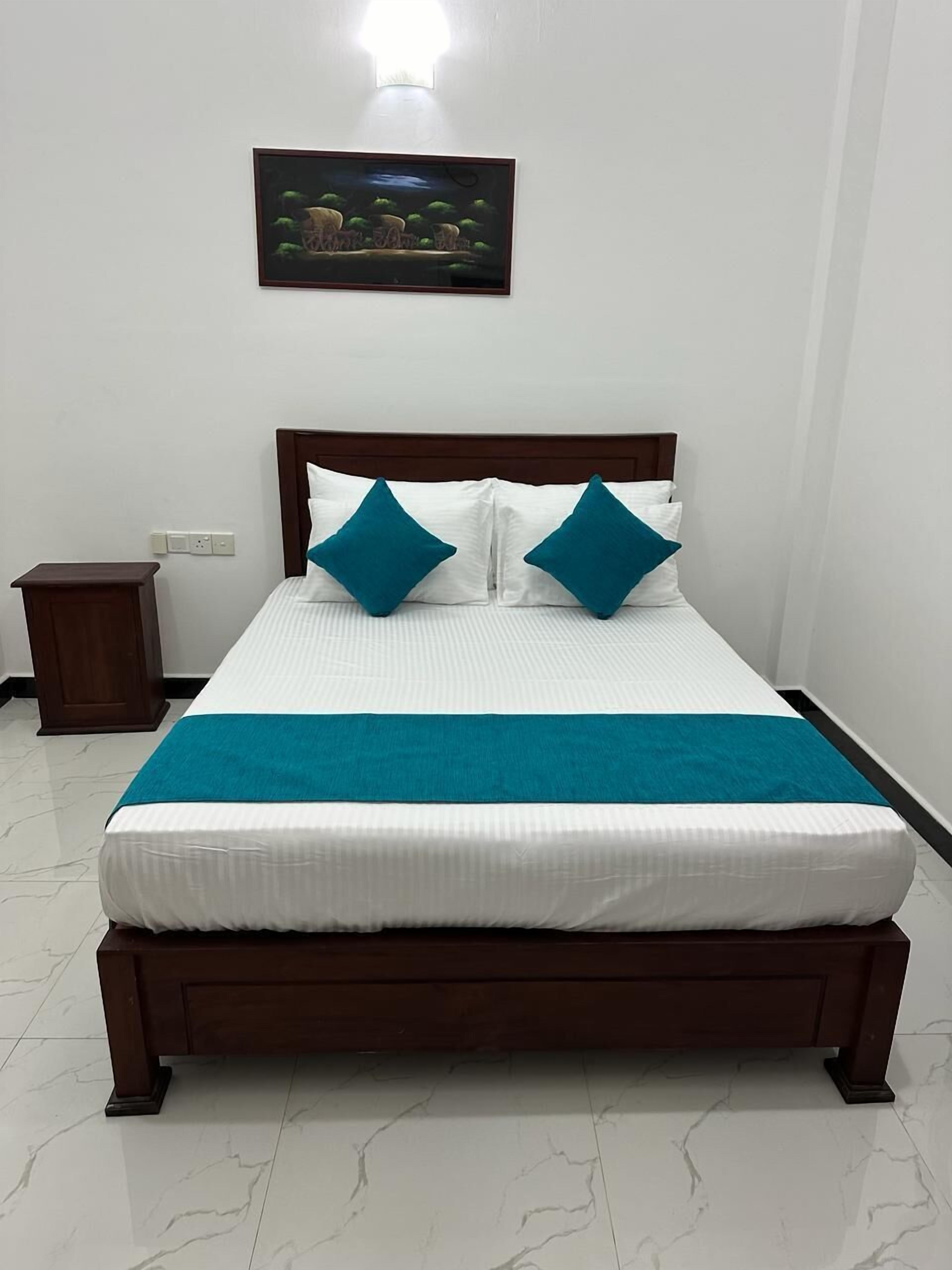 A neatly made bed with white bedding and two teal pillows, a wooden headboard, a small bedside table, and a wall-mounted light fixture.