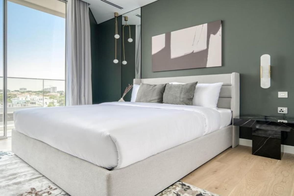 A modern bedroom with a large bed, a nightstand, a wall-mounted light, and a view through a large window.