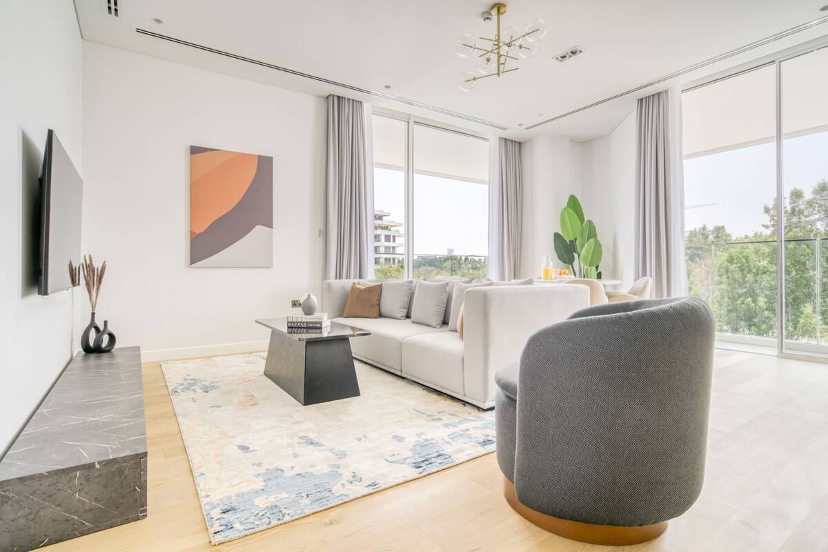 Una sala de estar moderna con un sofá, una mesa de centro y un sillón gris. Hay un ventanal con cortinas, un cuadro en la pared y una planta sobre una mesita de noche.