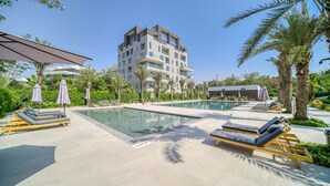 A pool area with lounge chairs and umbrellas, surrounded by palm trees and a modern building in the background. - Al Barari Stella Stays (Dubai)