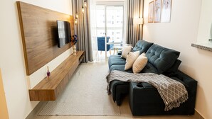 A modern living room with a black sofa, a mounted TV, a wooden bench, and a view of the outside through a large window. - Make Some Memories at This Unique Place (Praia Grande)