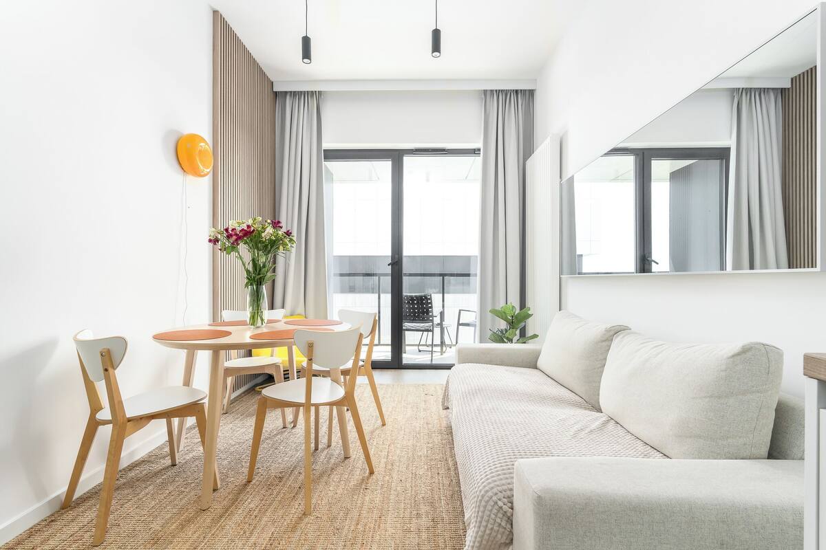 A modern living room with a sofa, a dining table with chairs, a large window, and a mirror.
