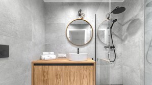 Un baño moderno con tocador de madera, un espejo redondo, un lavabo blanco y una zona de ducha con mampara de vidrio.