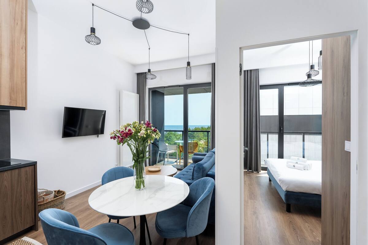 A modern hotel room with a balcony, a dining area with blue chairs, a round table, and a TV mounted on the wall.
