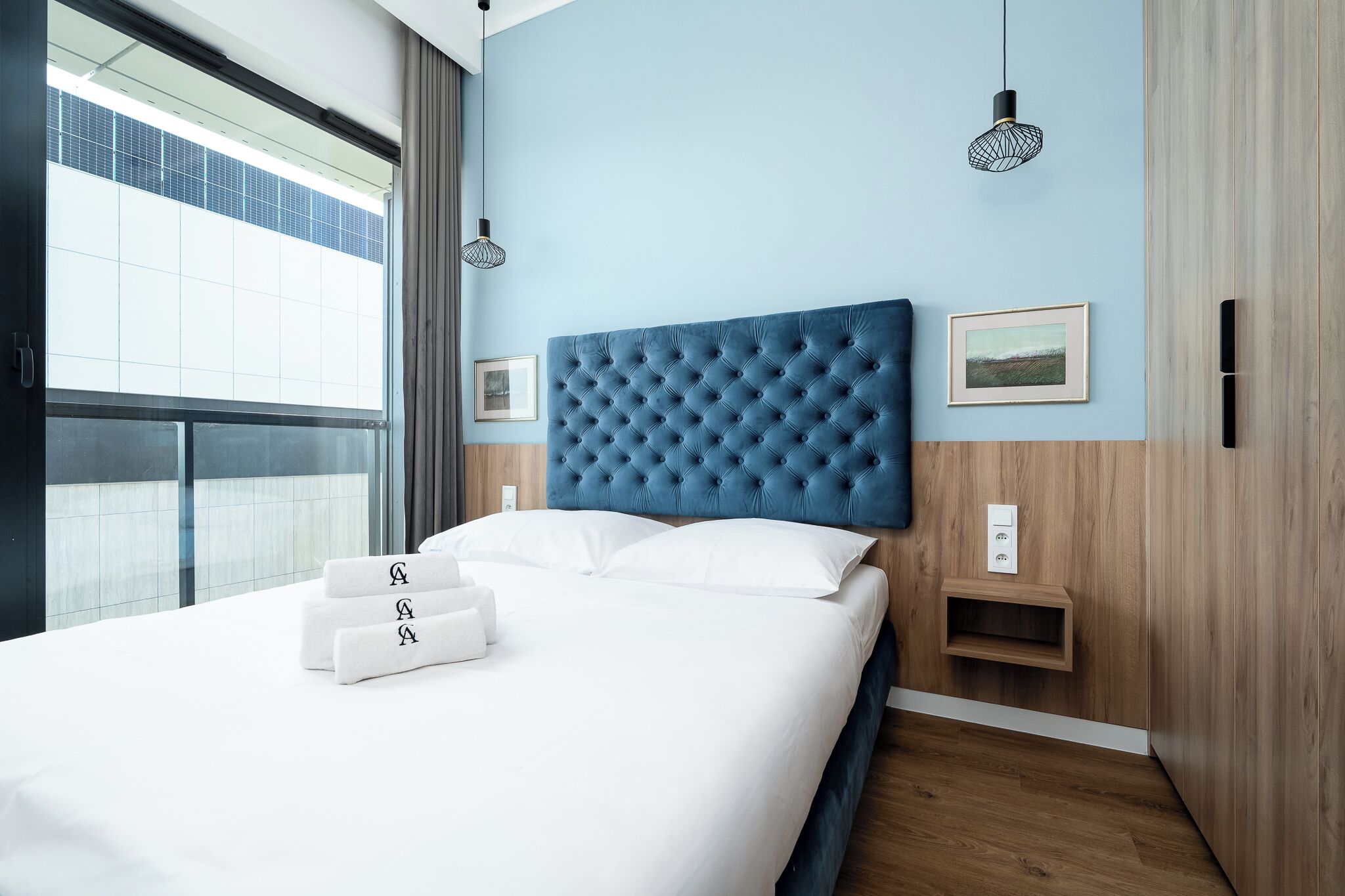 Una habitación de hotel moderna con una cama grande, cabecera azul y vistas amplias por una ventana grande.