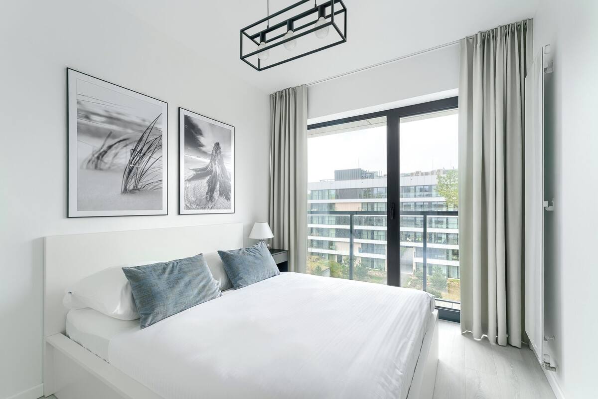 A modern hotel room with a large bed, two framed artworks, a bedside lamp, and a view of a cityscape through floor-to-ceiling windows.