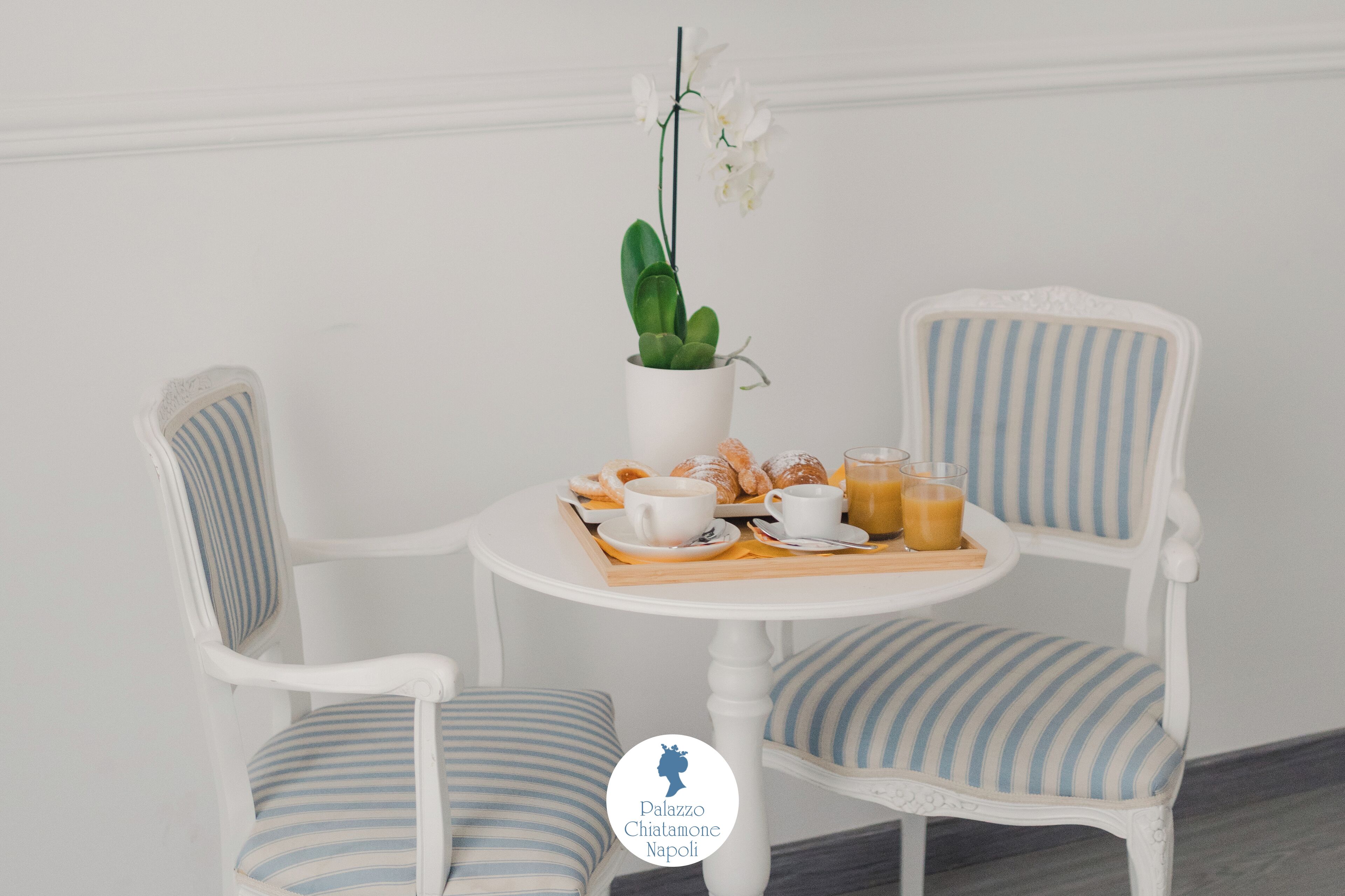 Uma mesa de jantar branca com cadeiras listradas, uma orquídea em vaso e itens de café da manhã, como croissants, café e suco de laranja.