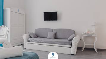 A modern bedroom with a grey sofa, a white wardrobe, a flat-screen TV, and a bedside table with a lamp.