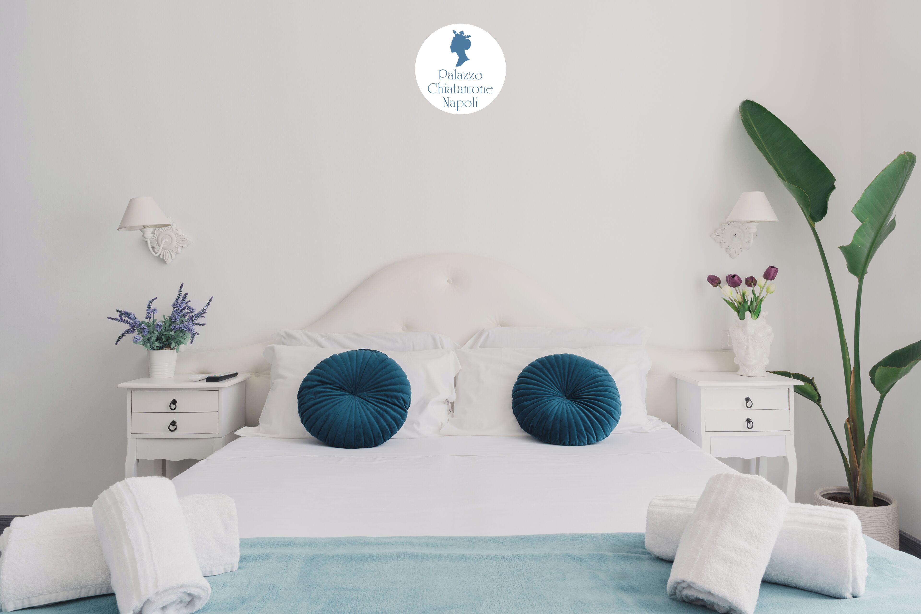 A neatly arranged bedroom with a white bed, two blue pillows, white bedside tables, and a potted plant.
