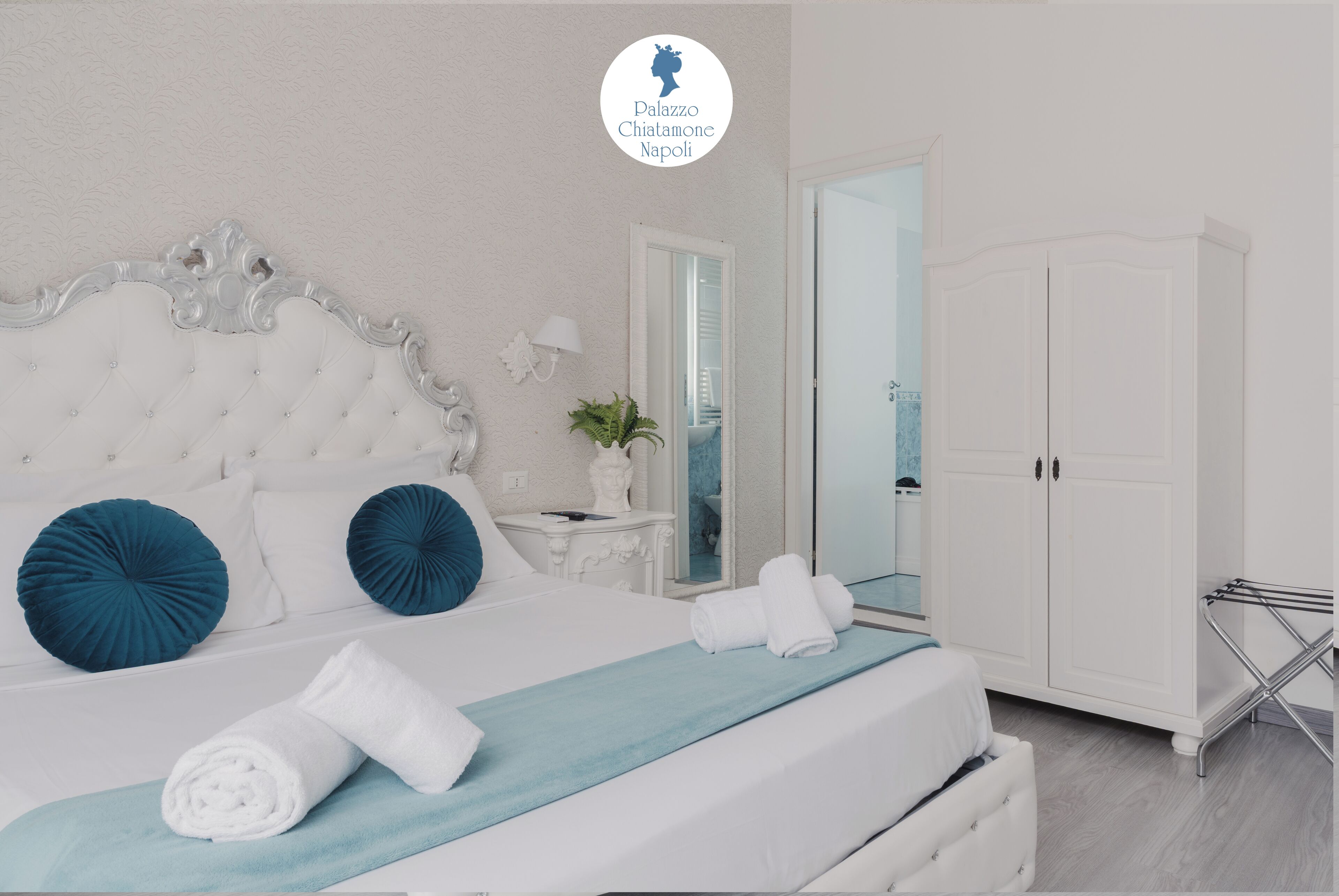 Un lit bien fait, doté d’une tête de lit capitonnée, de deux oreillers bleus et d’une couvre-lit bleue. On y trouve également une armoire blanche avec un miroir, une petite table avec une lampe et une plante.