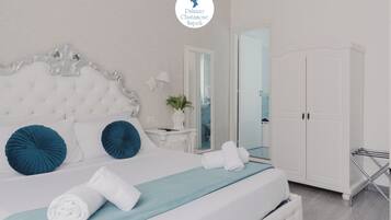 Un lit bien fait, doté d’une tête de lit capitonnée, de deux oreillers bleus et d’une couvre-lit bleue. On y trouve également une armoire blanche avec un miroir, une petite table avec une lampe et une plante.