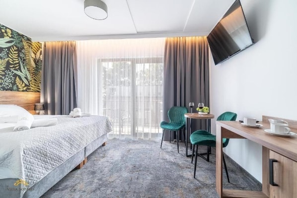 A hotel room with a bed, a desk with chairs, a TV, and a window with curtains. - Villa Solaris (Rewal)