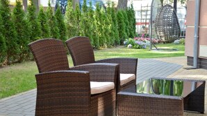 A wicker outdoor seating area with a glass table, surrounded by a well-maintained garden and a building in the background. - Villa Solaris (Rewal)