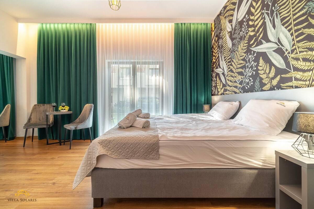 Ein modernes Schlafzimmer mit einem großen Bett, grünen Vorhängen, einem Essbereich mit Stühlen und einer Wand mit einem Blattmuster.
