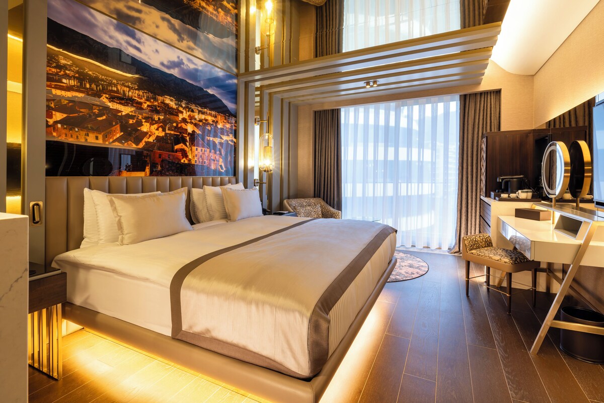 Ein modernes Hotelzimmer mit einem großen Bett, einem Schreibtisch mit Spiegel und einem Stadtbild-Wandgemälde.