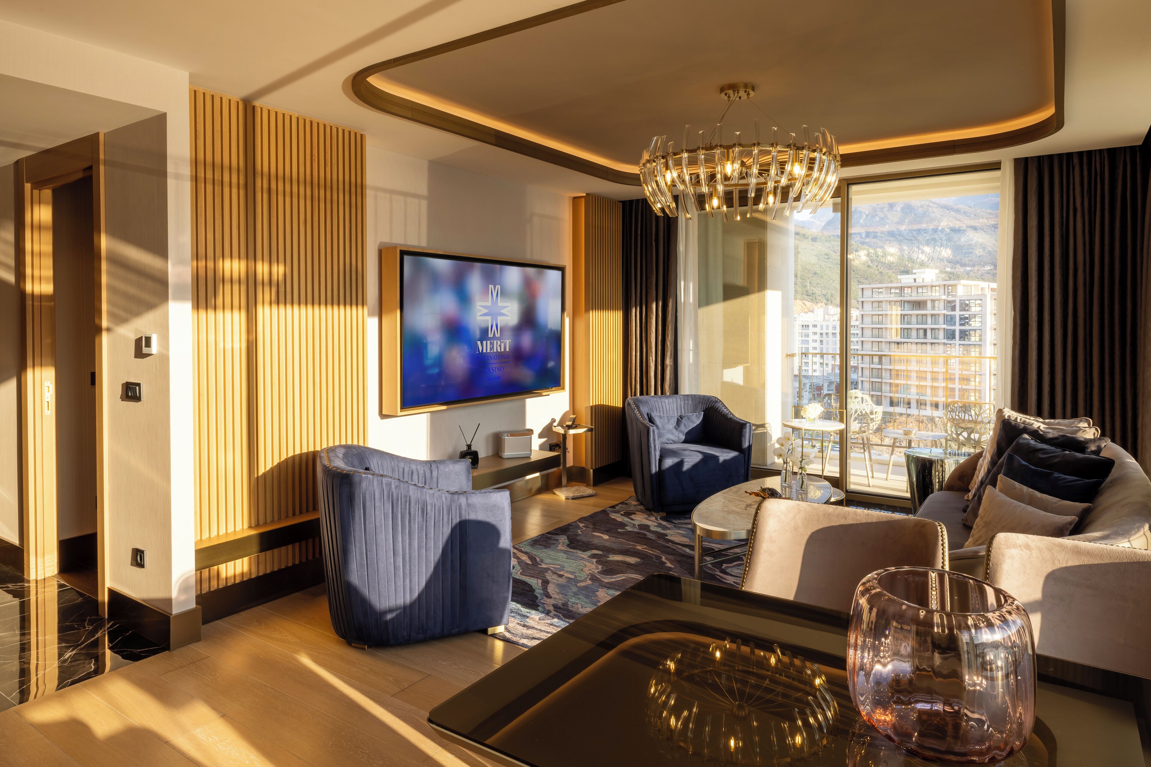 A modern hotel room with a large TV, comfortable seating, a chandelier, and a view of the city.