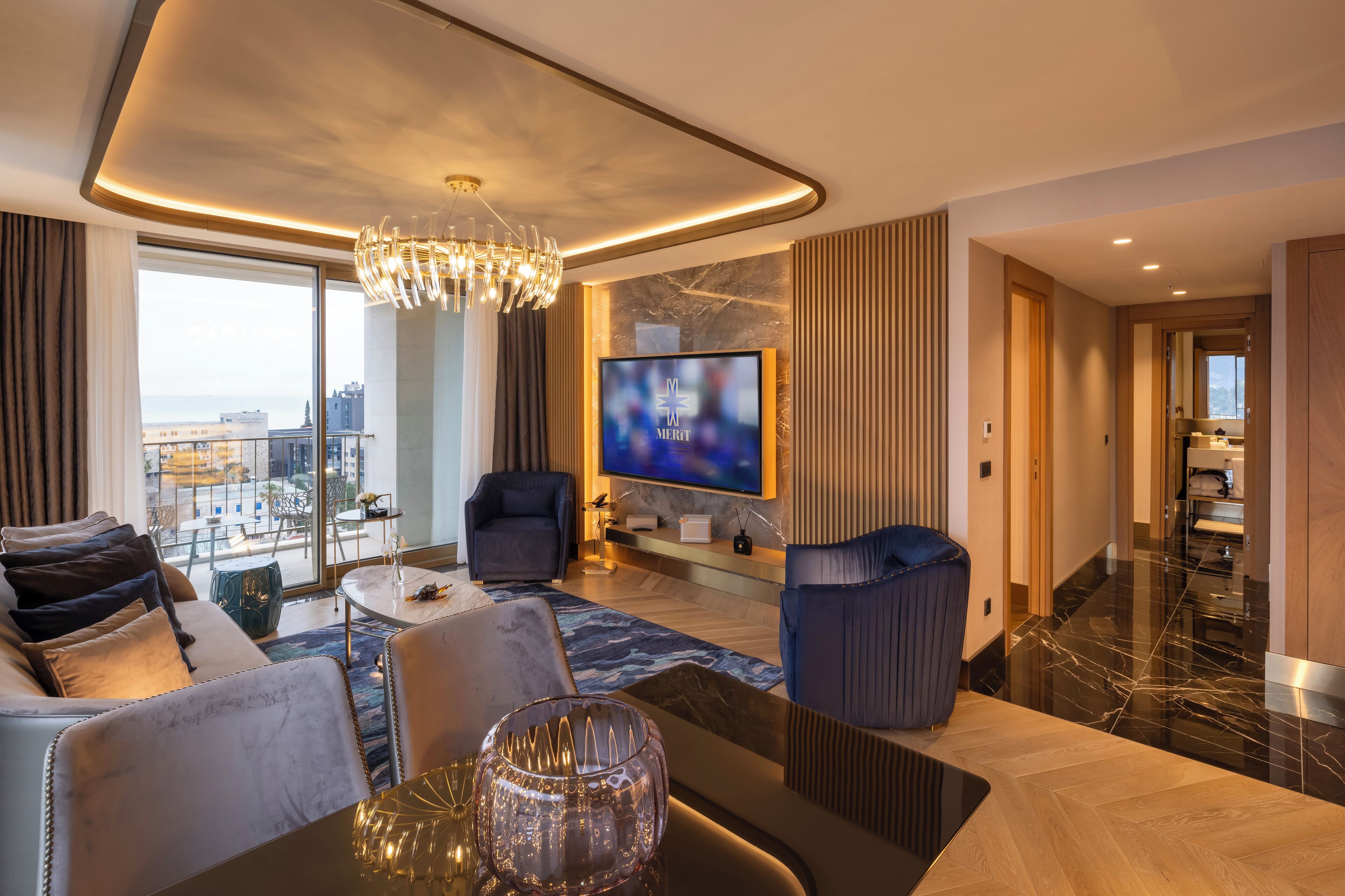 A modern living room with a large TV, comfortable seating, a chandelier, and a view of the city.